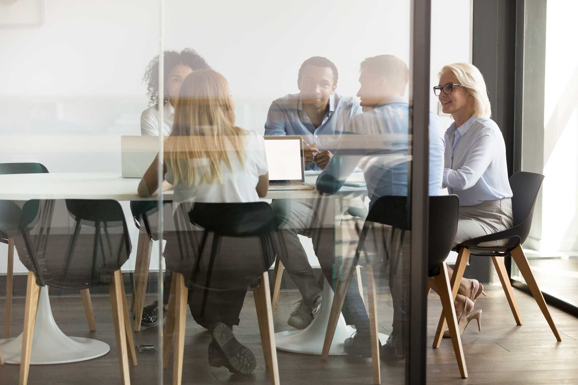 older woman leading meeting at conference table, distant, white, tan